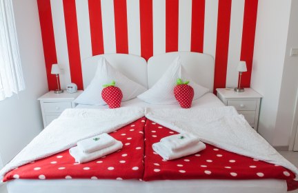 Doppelzimmer mit rot-weiß gestreifter Wand hinter dem Bett, roter Bettwäsche mit weißen Punkten und Erdbeerkissen